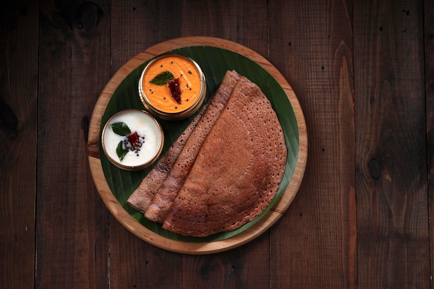 A crisp, thin, dark brown ragi dosa folded on a traditional steel plate, served with a small bowl of fresh white coconut chutney on the side.